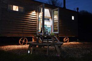 Meidrim Meadows shepherd's hut at night with the doors open and the lights on inside. Outside a table can be seen in the foreground with evening drinks set up.