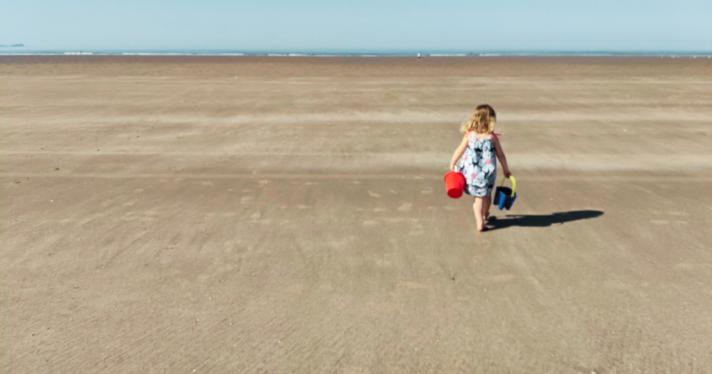 A child dressed in a sun dress is walking towards the sea on the vast golden sand on Pembrey Beach. She is holding two buckets. It is completely secluded.