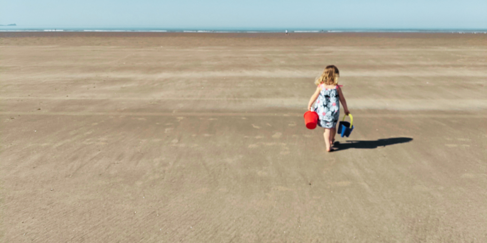 A child dressed in a sun dress is walking towards the sea on the vast golden sand on Pembrey Beach. She is holding two buckets. It is completely secluded.