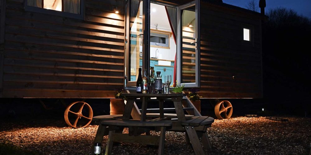 Meidrim Meadows shepherd's hut at night with the doors open and the lights on inside. Outside a table can be seen in the foreground with evening drinks set up.